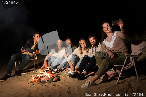 Image of happy friends taking selfie at camp fire on beach