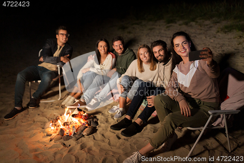 Image of happy friends taking selfie at camp fire on beach