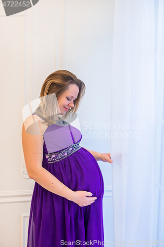 Image of Beautiful pregnant woman in ultra violet dress standing near window