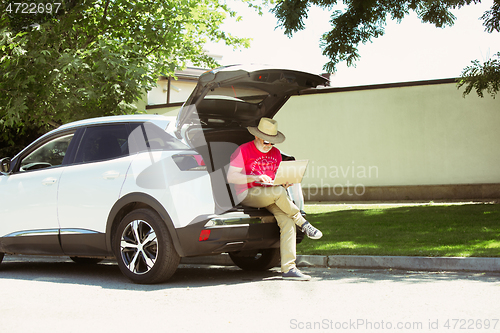 Image of Senior man sitting on his car\'s trunk and working outdoors