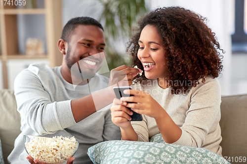 Image of african couple with popcorn and smartphone at home