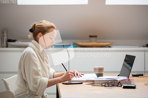 Image of Young female entrepreneur wearing cosy warm bathrobe working remotly from home in winter Christmas time during 2020 corona virus pandemic. Work from home, selter in place, concept.