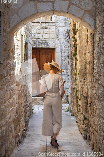 Image of Rear view of beautiful blonde young female traveler wearing straw sun hat sightseeing and enjoying summer vacation in an old traditional costal town at Adriatic cost, Croatia