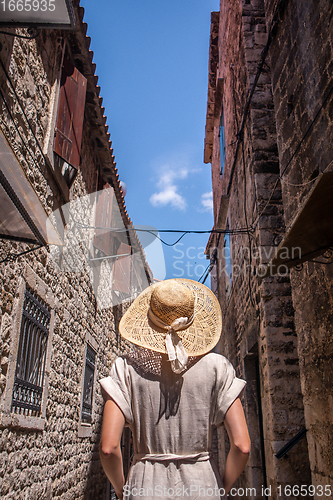 Image of Rear view of beautiful blonde young female traveler wearing straw sun hat sightseeing and enjoying summer vacation in an old traditional costal town at Adriatic cost, Croatia