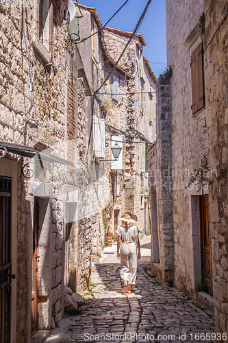 Image of Rear view of beautiful blonde young female traveler wearing straw sun hat sightseeing and enjoying summer vacation in an old traditional costal town at Adriatic cost, Croatia