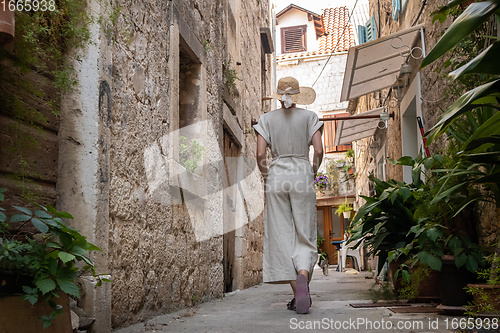 Image of Rear view of beautiful blonde young female traveler wearing straw sun hat sightseeing and enjoying summer vacation in an old traditional costal town at Adriatic cost, Croatia