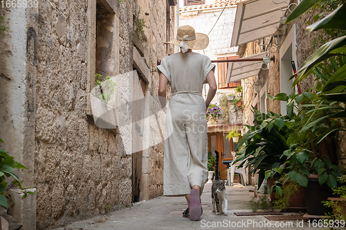 Image of Rear view of beautiful blonde young female traveler wearing straw sun hat sightseeing and enjoying summer vacation in an old traditional costal town at Adriatic cost, Croatia