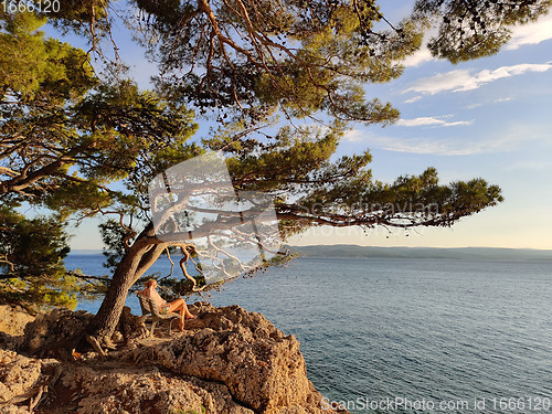 Image of Pensive woman on vacations, sitting and relaxing under large pine tree on bench by dip blue sea enjoying beautiful sunset light in Brela, Makarska region, Dalmatia, Croatia