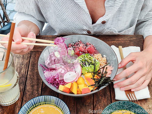 Image of Woman eating tasty colorful healthy natural organic vegetarian Hawaiian poke bowl using asian chopsticks on rustic wooden table. Healthy natural organic eating concept