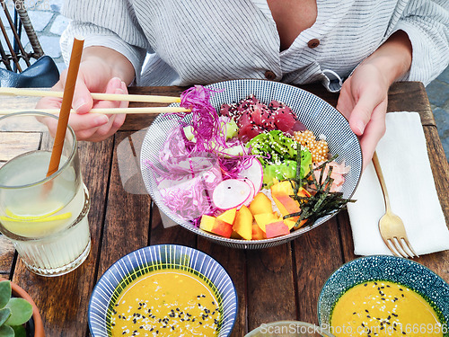 Image of Woman eating tasty colorful healthy natural organic vegetarian Hawaiian poke bowl using asian chopsticks on rustic wooden table. Healthy natural organic eating concept