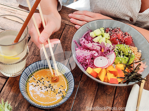 Image of Woman eating tasty colorful healthy natural organic vegetarian Hawaiian poke bowl using asian chopsticks on rustic wooden table. Healthy natural organic eating concept