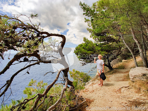Image of Young active feamle tourist taking a break, drinking water, wearing small backpack while walking on coastal path among pine trees looking for remote cove to swim alone in peace on seaside in Croatia