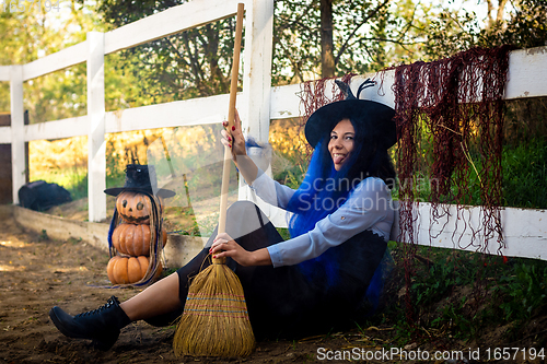 Image of A girl dressed as a witch sits by a fence with a broom in her hands and shows her tongue