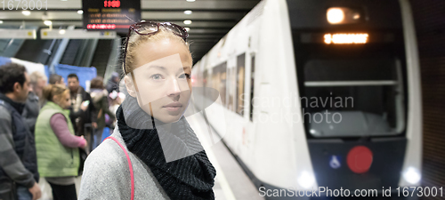 Image of Young woman in winter coat waiting on the platform of a railway station for train to arrive. Public transport