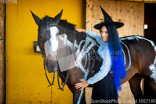 Image of A girl dressed as a witch stands by a horse on which a skeleton is painted in white paint