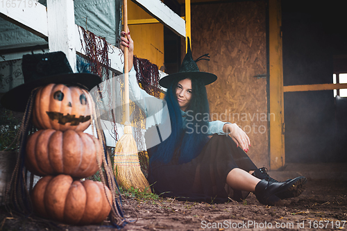 Image of A girl dressed as a witch sits by a fence with a broom in her hands, in the foreground there is a scary figure of pumpkins