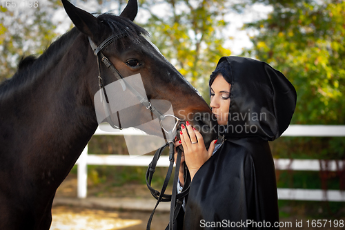 Image of Girl in a black cloak hugs and kisses a horse