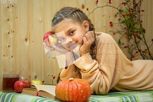 Image of A girl lying down reads a book, a pumpkin and apples lie nearby, a girl joyfully looks into the frame