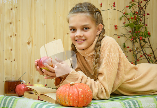 Image of A girl lying down reads a book, holds an apple in her hands and looks happily into the frame