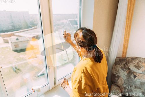 Image of Woman Of Fifty In Yellow Sweater And Jeans Washes Dusty Window In Apartment. 50 Year Old Woman Cleans Windows From Stains Using Rag And Spray Cleaner. Caucasian Elderly Woman Is Cleaning House, Doing