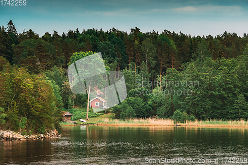 Image of Sweden. Beautiful Red Swedish Wooden Log Cabin House On Rocky Island Coast In Summer. Lake Or River And Forest Landscape