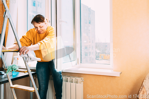 Image of Smiling Caucasian Woman Of Fifty In Yellow Sweater And Jeans Washes Dusty Window In Apartment. 50 Year Old Woman Cleans Windows From Stains Using Rag And Spray Cleaner. Elderly Woman Is Cleaning Hous