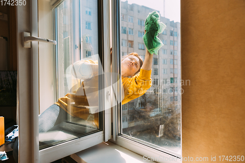 Image of Caucasian Woman Of Fifty In A Yellow Sweater And Jeans Washes A Dusty Window In Apartment. A 50 Year Old Woman Cleans Windows From Stains Using Rag. Elderly Woman Is Cleaning House, Doing Household C