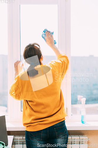 Image of Woman Of Fifty In Yellow Sweater And Jeans Washes Dusty Window In Apartment. 50 Year Old Woman Cleans Windows From Stains Using Rag And Spray Cleaner. Caucasian Elderly Woman Is Cleaning House, Doing