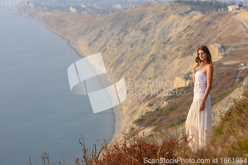 Image of A beautiful slender girl in a white dress stands on a hill and enjoys a beautiful view of the sunset