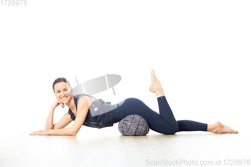 Image of Restorative yoga with a bolster. Young sporty female yoga instructor in bright white yoga studio, lying on bolster cushion, stretching, smilling, showing love and passion for restorative yoga