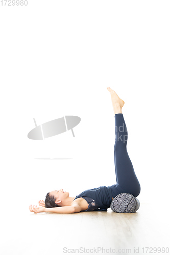 Image of Restorative yoga with a bolster. Young sporty attractive woman in bright white yoga studio, lying on bolster cushion, stretching and relaxing during restorative yoga. Healthy active lifestyle