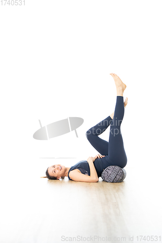 Image of Restorative yoga with a bolster. Young sporty attractive woman in bright white yoga studio, lying on bolster cushion, stretching and relaxing during restorative yoga. Healthy active lifestyle