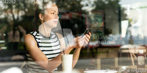 Image of Thoughtful caucasian woman holding mobile phone while looking through the coffee shop window during coffee break.