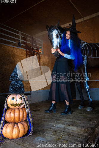 Image of A girl in a witch costume hugs a horse in a corral, in the foreground an evil figure of pumpkins