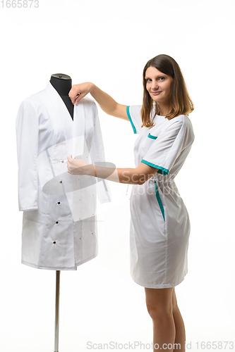 Image of A medical gown hangs on a mannequin, a girl nurse straightens the collar of a gown