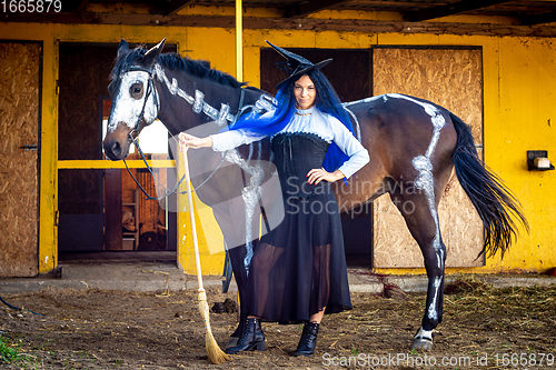 Image of A girl dressed as a witch stands with a broom near a horse on which a skeleton is drawn