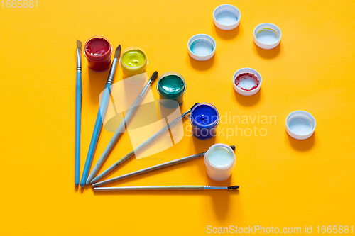 Image of On a yellow background, brushes and jars of gouache are laid out, lids are randomly laid out