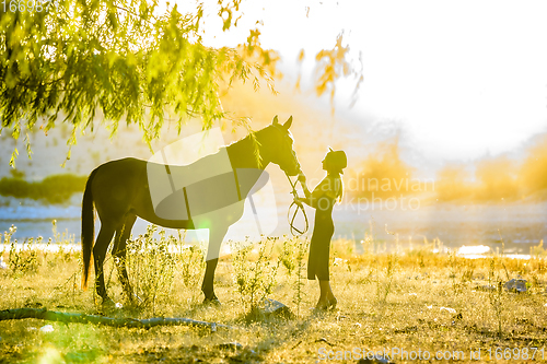 Image of A girl stands in front of a horse on the shore of a lake and is flooded with warm sunbeams at sunset