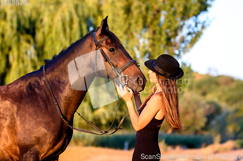 Image of A girl in a black hat in the rays of the setting sun looks happily at a horse