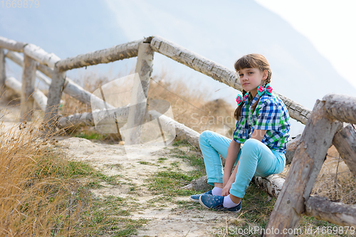 Image of A girl sits on a fence made of a wooden blockhouse against the background of a landscape