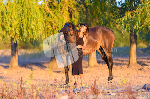 Image of A girl with a horse stands against the background of trees, the rays of the setting sun fall on them