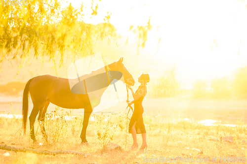 Image of A girl stands in front of a horse, they are bathed in warm sunbeams at sunset