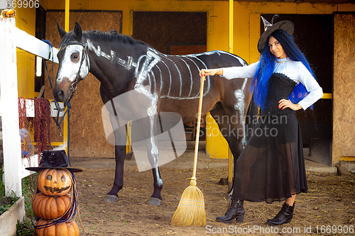 Image of A witch on a farm stands next to a horse and an evil pumpkin man