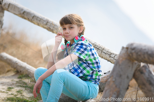 Image of A girl sits on a fence made of a wooden blockhouse and, with a blade of grass in her mouth, looks into the frame