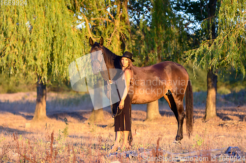 Image of A slender beautiful girl in a black dress and hat walks with a horse