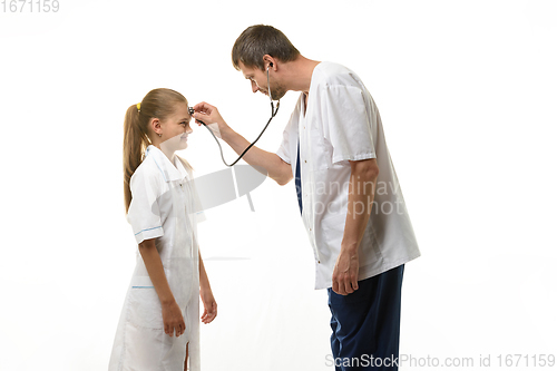 Image of The doctor put the head of the phonendoscope to the forehead of the girl in a medical gown