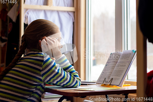 Image of The schoolgirl carefully reads the task in the textbook while sitting at the table by the window at home