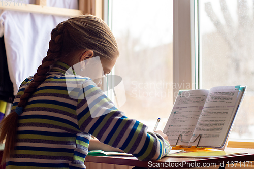 Image of Schoolgirl sits at a table by the window and does her homework, view from the back