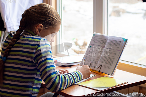 Image of Schoolgirl sits at the table by the window and does homework