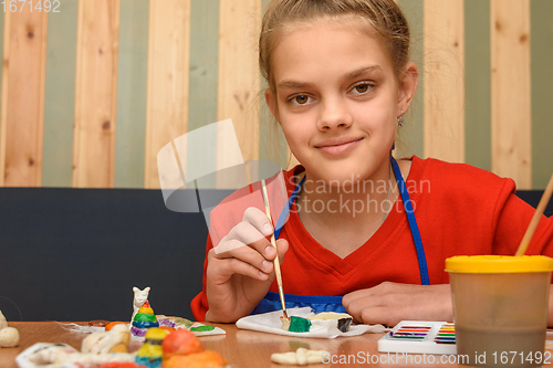 Image of A girl paints a craft from salt dough with watercolors and looked into the frame